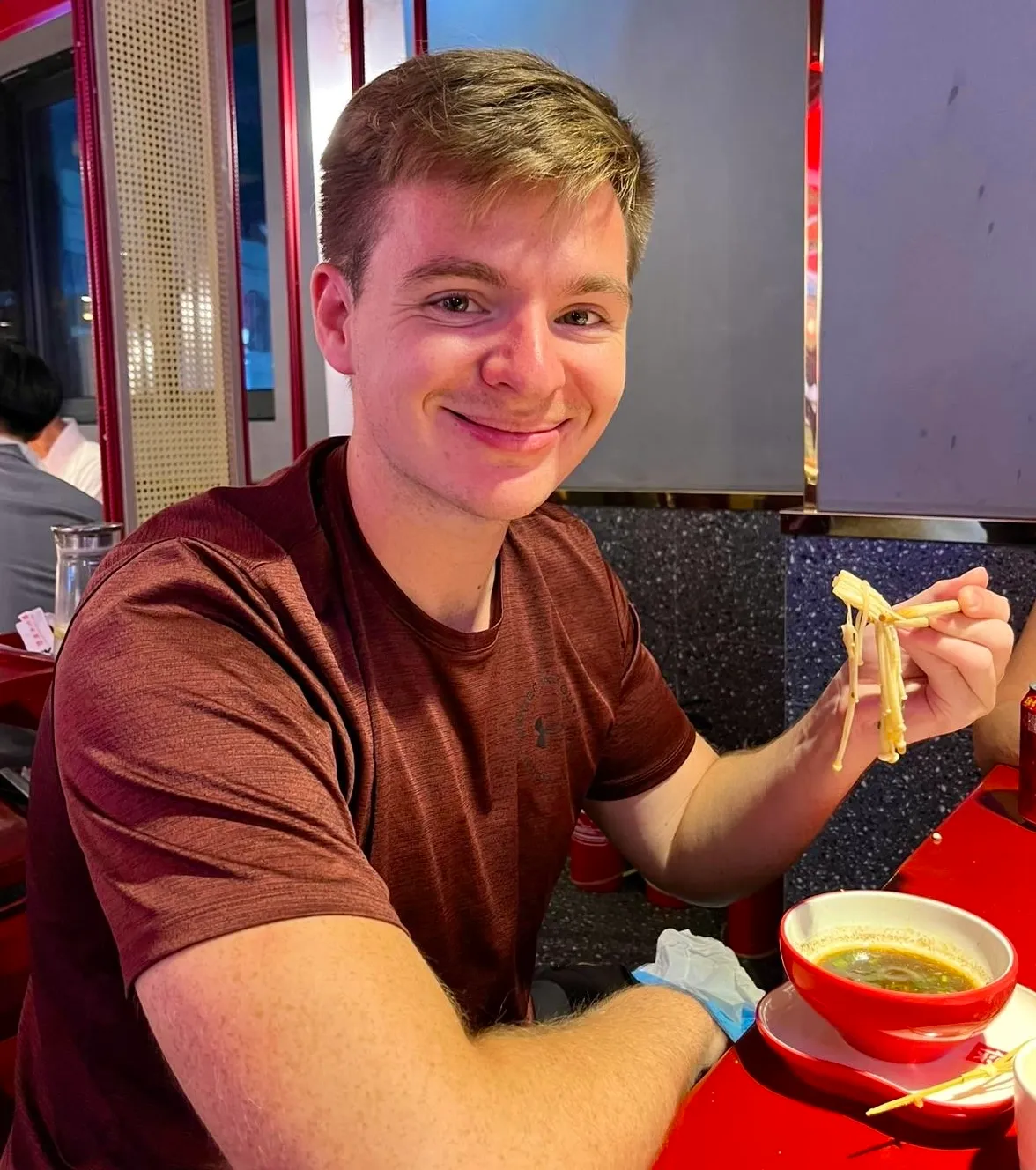 Niklas mit Stäbchen und einer Schale Nudelsuppe in einem Lokal in China, Neonlicht und rote Tische